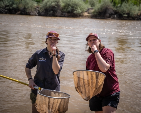 Two students holding razorback sucker and kissing them goodbye. The students are holding fishing nets and standing in the Colorado River.